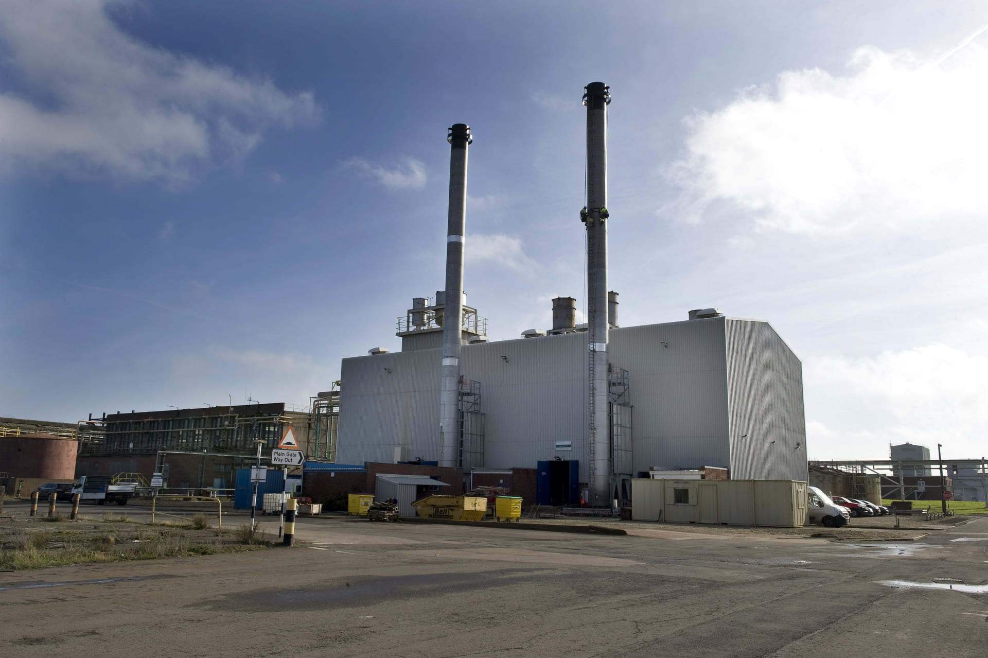 Stallingborough, an industrial facility with two tall chimneys standing side by side, likely part of a power plant or manufacturing site. The building has a boxy, utilitarian design and is surrounded by an open yard with scattered containers and equipment. The sky is partly cloudy, casting a mix of shadows and sunlight over the scene.