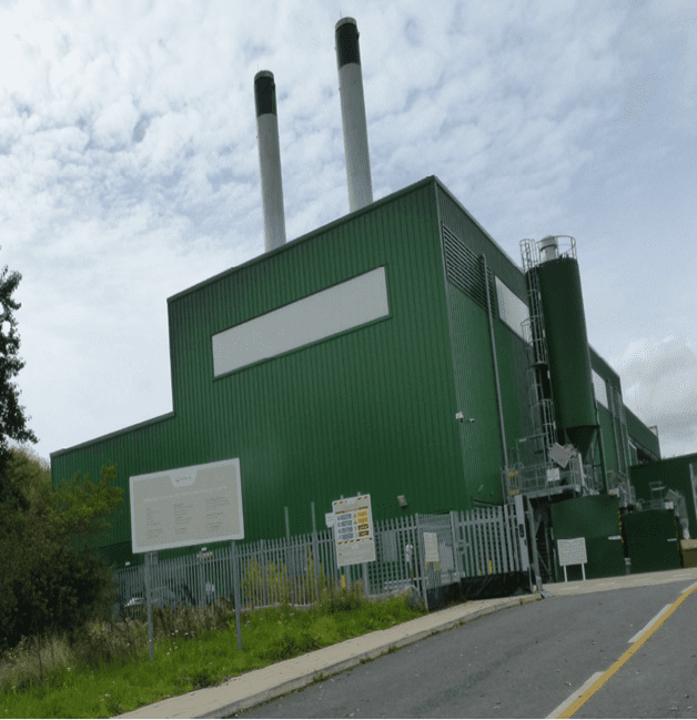 The image is the Holbrook biomass CHP facility in Sheffield. The surroundings include a road, trees, a building, grass and the sky is visible with clouds.