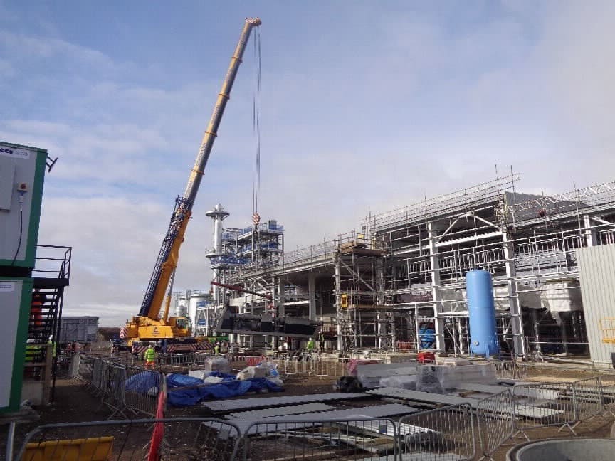 The image is of a crane at a construction site in Wilton ELP. It shows the crane against the sky, with outdoor industrial engineering and construction activities in progress. The scene includes steel pipes and other construction equipment. Plastic recycling plant.
