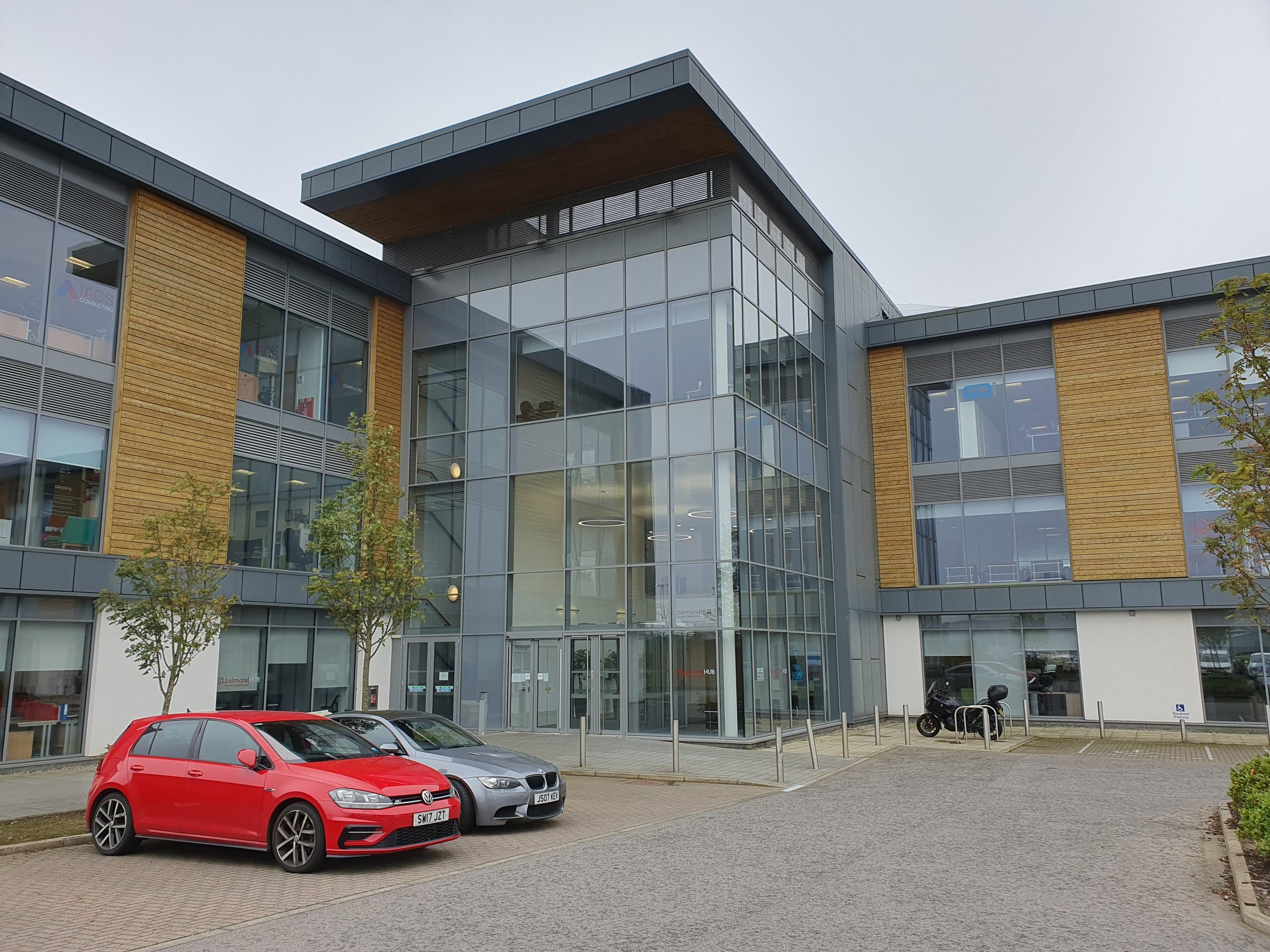 Modern office building with large glass windows and wood-paneled accents, featuring a central entrance and surrounded by parked vehicles and minimal landscaping. Aberdeen Office | px Group
