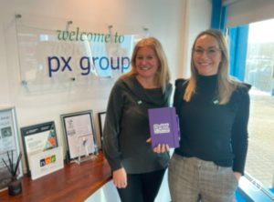 Two women employees standing in the px office reception holding a power of women book in efforts to support women’s empowerment.